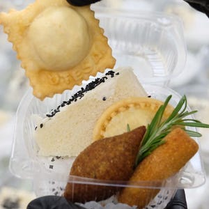 Puede incluir: Un recipiente de pl&aacute;stico transparente lleno de varios aperitivos. Incluye un ravioli frito, un s&aacute;ndwich con semillas de s&eacute;samo negro, una tarta peque&ntilde;a, una alb&oacute;ndiga frita y un rollo frito, adornado con romero.