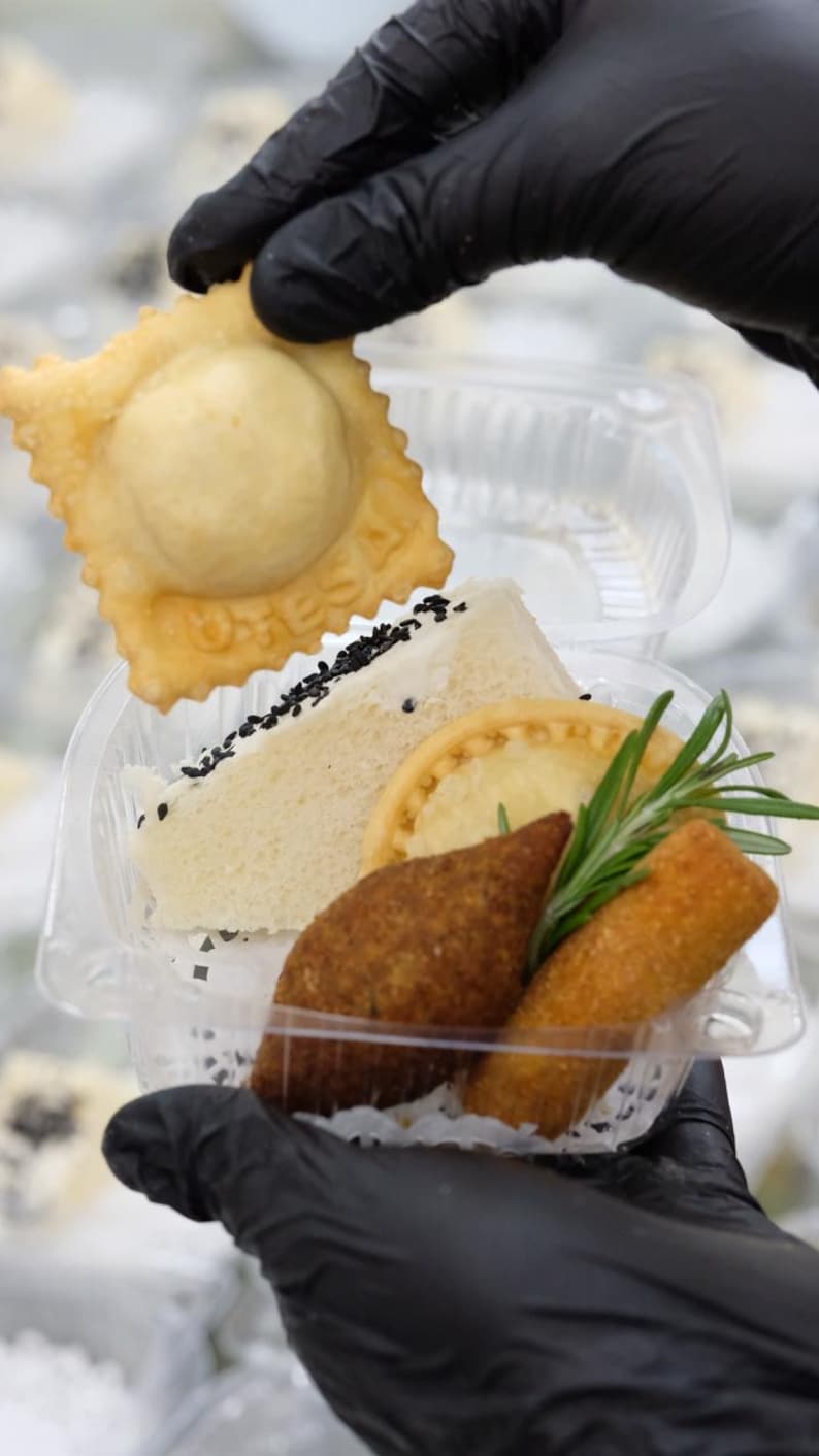Puede incluir: Un recipiente de pl&aacute;stico transparente lleno de varios aperitivos. Incluye un ravioli frito, un s&aacute;ndwich con semillas de s&eacute;samo negro, una tarta peque&ntilde;a, una alb&oacute;ndiga frita y un rollo frito, adornado con romero.