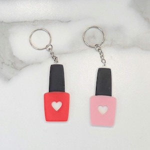Op de afbeelding: Twee sleutelhangers in de vorm van nagellak flesjes, een rode en een roze, met een witte hartvormige uitsparing op elk. De sleutelhangers hebben zilveren sleutelringen.