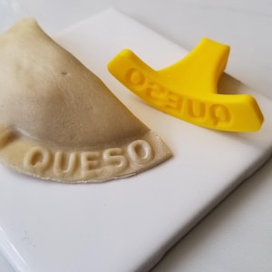 Puede incluir: Una empanada beige con la palabra "QUESO" grabada en la corteza, junto a una herramienta de plástico amarilla con la palabra "ONE20" grabada. Los artículos están sobre una baldosa cuadrada blanca.