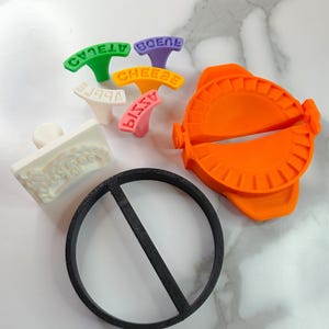 Puede incluir: Una colecci&oacute;n de herramientas de cocina coloridas, que incluyen un fabricante de ravioles de color naranja brillante, un cortador circular negro y varios sellos de alimentos con texto. Los sellos son verdes, morados, amarillos, blancos y rosas, cada uno con una palabra diferente.