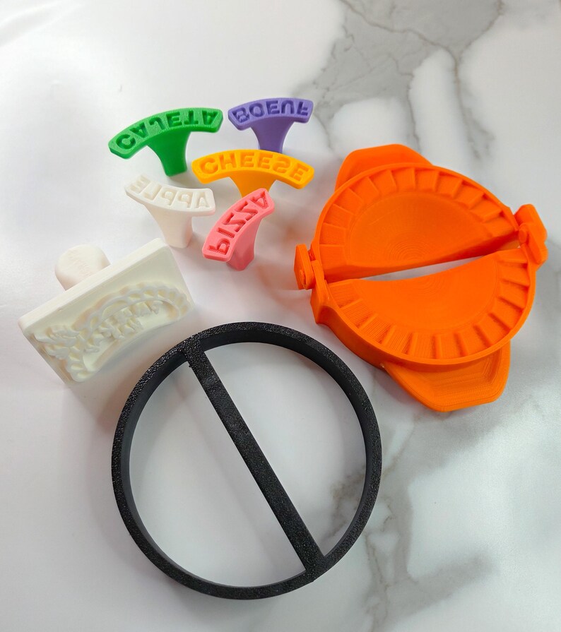 Puede incluir: Una colecci&oacute;n de herramientas de cocina coloridas, que incluyen un fabricante de ravioles de color naranja brillante, un cortador circular negro y varios sellos de alimentos con texto. Los sellos son verdes, morados, amarillos, blancos y rosas, cada uno con una palabra diferente.