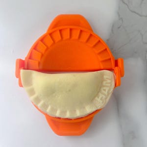 May include: An orange dumpling maker with a semi-circular design, holding a light-colored dumpling. The dumpling maker has a ridged edge and a handle. The dumpling has the word "HAM" imprinted on it.
