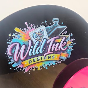 Op de afbeelding: Een rond zwart bord met een kleurrijk logo en de tekst Wild Ink DESIGNS. De afbeelding bevat een hart met een luipaardprint, een hittepers en levendige verfspatten in de kleuren blauw, roze en geel.