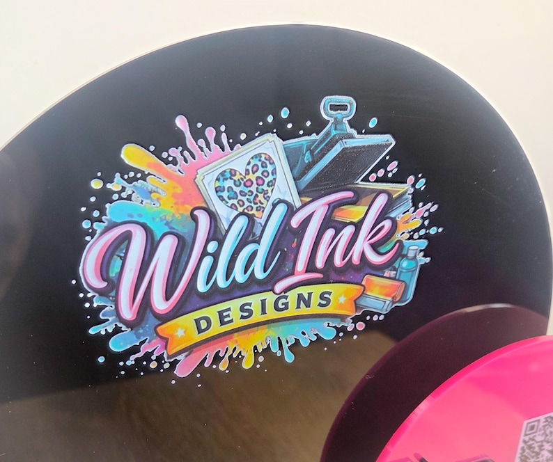 Op de afbeelding: Een rond zwart bord met een kleurrijk logo en de tekst Wild Ink DESIGNS. De afbeelding bevat een hart met een luipaardprint, een hittepers en levendige verfspatten in de kleuren blauw, roze en geel.