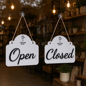 Peut inclure: Deux panneaux blancs en forme de nuage suspendus par de la ficelle à des ventouses sur une porte vitrée. Le panneau de gauche porte le texte noir "Open" et le logo "Lux Lab Studio". Celui de droite porte le texte "Closed" et le même logo.
