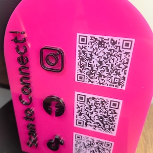 Op de afbeelding: Een felroze, boogvormig acrylbord met de zwarte tekst "Scan to Connect!" verticaal aan de linkerkant. Het bevat drie verhoogde zwarte social media iconen en drie bijbehorende witte QR-codes die aan de rechterkant zijn afgedrukt.