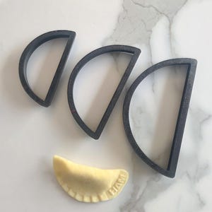 May include: Three black, semi-circular cookie cutters of varying sizes are arranged above a single, filled pierogi. The pierogi is light yellow with a crimped edge and the word "HAM" stamped on it.