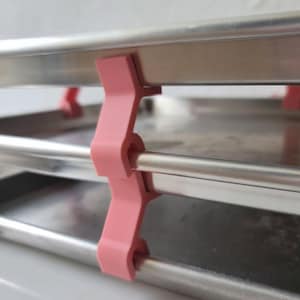May include: A close-up shot of a pink plastic clip fastening two metal rods and a metal tray. The clip is a pale pink colour and has an unusual shape. The rods and tray are made of a shiny, silver metal. The background is a plain white surface.