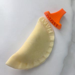 Puede incluir: Un pierogi crudo con la palabra "HAM" impresa, junto a un sello de pierogi naranja. El pierogi es de color amarillo claro y el sello es de color naranja brillante.