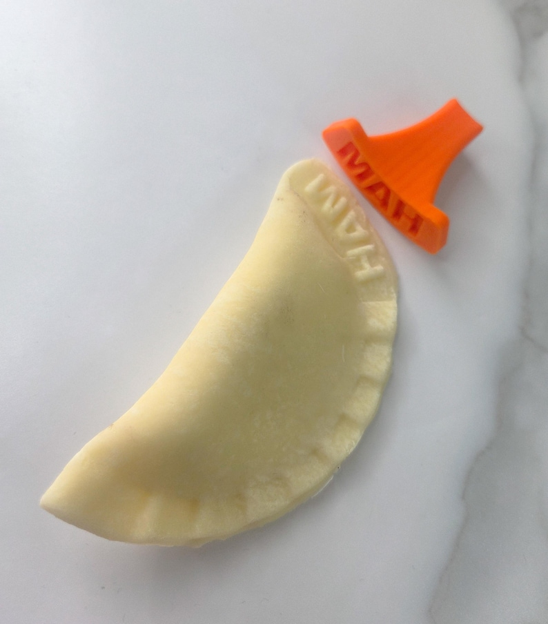 Puede incluir: Un pierogi crudo con la palabra "HAM" impresa, junto a un sello de pierogi naranja. El pierogi es de color amarillo claro y el sello es de color naranja brillante.