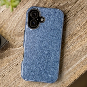 Peut inclure: Un étui de téléphone bleu avec une texture denim et une découpe pour un appareil photo à double objectif. L'étui est posé sur une surface en bois, avec un appareil photo et de la verdure en arrière-plan. L'étui est conçu pour protéger le téléphone.