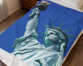 Puede incluir: Una manta de forro polar suave con un estampado grande y detallado de la Estatua de la Libertad en verde claro sobre un fondo azul sólido. El diseño muestra la estatua desde el pecho hacia arriba, sosteniendo una antorcha con llama dorada.