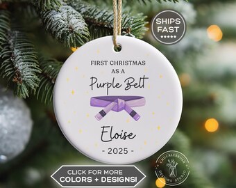 May include: A round white ceramic ornament hanging from a tan string. The text reads FIRST CHRISTMAS AS A Purple Belt, Eloise, and 2025. A graphic of a tied purple martial arts belt is centered between the text, surrounded by small yellow stars.