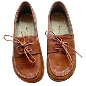Könnte beinhalten: Ein Paar braune Lederschnürschuhe mit einem Steppdetail am Rand. Die Schuhe haben eine braune Lederschnur und der Markenname "SWISS" ist auf der Innenseite jedes Schuhs aufgedruckt.