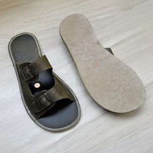 May include: A pair of dark brown leather sandals with two adjustable buckle straps. The sandals have a contoured footbed and a stitched edge. The sole is a light tan color. The sandals are displayed on a light wood surface.