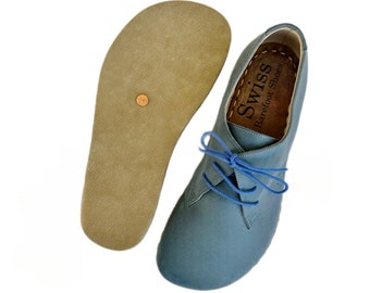 May include: A pair of blue suede lace-up shoes with a brown leather insole. The shoes have a round toe and a low heel. The insole is labeled "Swiss Barefoot Shoes".