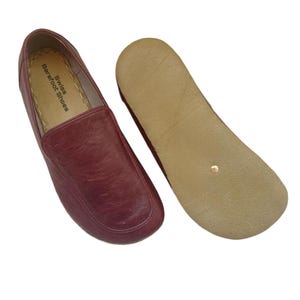 Pode incluir: Uma caneca de cerâmica branca com uma alça preta e o texto "But First, Coffee" em negrito preto. A caneca está sobre um fundo desfocado, sugerindo um ambiente acolhedor.