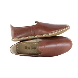 Puede incluir: Un par de zapatos marrones de cuero sin cordones con una costura cosida alrededor de la suela. Los zapatos están apilados uno encima del otro y la plantilla del zapato superior tiene el texto "Swiss Handmade Shoes".