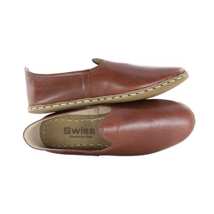 Könnte beinhalten: Zwei Paar braune Lederschlappen mit weißen Nähten an der Sohle. Die Schuhe sind übereinander gestapelt. Die Innensohle des oberen Schuhs trägt den Text "Swiss Handmade Shoes".