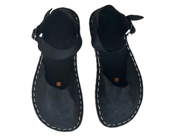 Puede incluir: Un par de sandalias de cuero negro con diseño de punta redondeada, una correa de tobillo con hebilla de metal y costuras blancas en contraste a lo largo del borde exterior de la suela. Un pequeño detalle circular marrón es visible en la plantilla.
