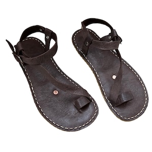 May include: A pair of brown leather sandals with ankle straps and a toe loop. The sandals have a simple design and are stitched with white thread.