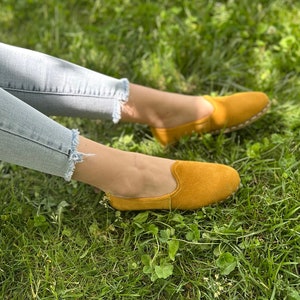 Pode incluir: Um par de mocassins de camurça amarelos com bico fino. Os mocassins são usados por uma pessoa sentada em uma superfície gramada.