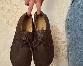 Op de afbeelding: Een paar bruine leren schoenen met veters. De schoenen worden vastgehouden door de vingers van een persoon. De schoenen hebben een ronde neus en een gestikte zool. De achtergrond is een behang met patroon.