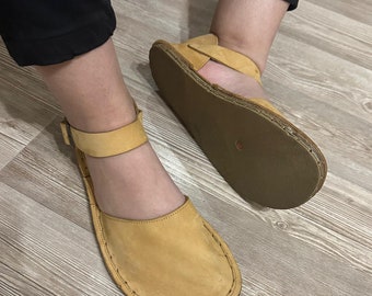 May include: A pair of tan leather sandals with a single strap and buckle closure. The sandals have a stitched edge and a flat sole.