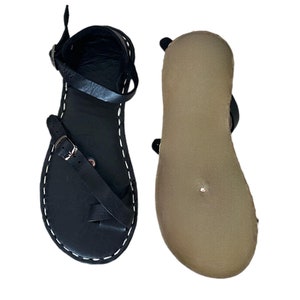 Peut inclure: Une paire de sandales en cuir noir avec une seule sangle sur le pied et une boucle de fermeture réglable. Les sandales ont une semelle intérieure en cuir marron et un bord cousu.