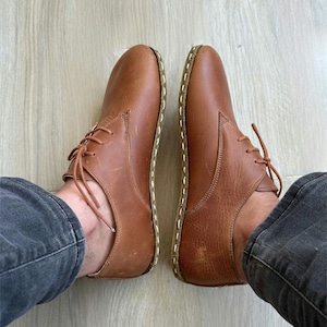 以下が含まれることがあります： 縁に白いステッチが施された、茶色の革製のレースアップシューズのペア。暗いデニムのジーンズを履いた人が履いています。