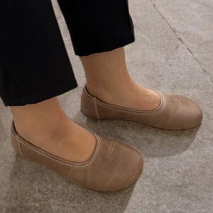 Op de afbeelding: Een paar lichtbruine ballerina's. De schoenen zijn gemaakt van een zacht materiaal en hebben een ronde neus. De schoenen worden gedragen met een zwarte broek. De schoenen hebben een neutrale kleur.