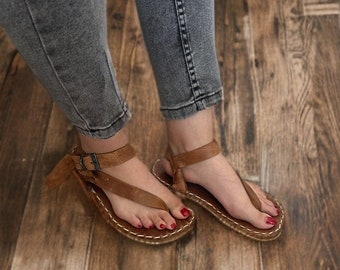 Può includere: Un paio di sandali in pelle marrone con cinturino con fibbia e suola intrecciata. I sandali sono indossati su un pavimento in legno.