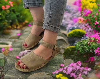 May include: A pair of brown leather sandals with ankle straps. The sandals are worn by a person standing on a stone path surrounded by flowers.