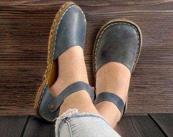 May include: A pair of slate blue leather flats featuring a rounded toe, an ankle strap with a small metallic button closure, and a visible jute-wrapped sole edge with thick white contrast stitching along the perimeter of the shoe base.