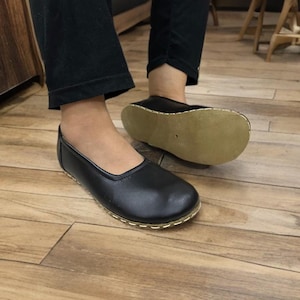 May include: Black leather slip-on shoes with a rounded toe and a gold-colored stitched trim. The soles are a light tan color. The shoes are on a wooden floor.