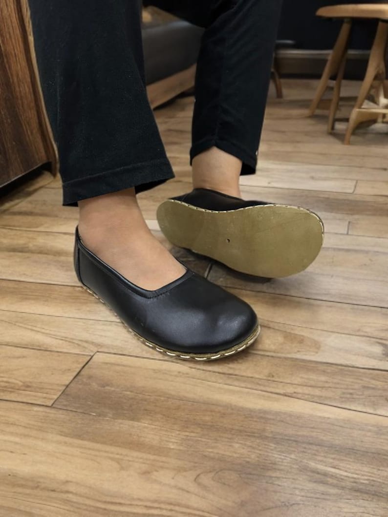 May include: Black leather slip-on shoes with a rounded toe and a gold-colored stitched trim. The soles are a light tan color. The shoes are on a wooden floor.