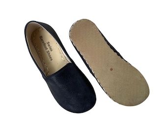 Può includere: Un paio di scarpe slip-on in pelle nera con punta arrotondata. Una scarpa è a testa in su e l'altra a testa in giù, rivelando una suola color beige. L'interno della scarpa riporta la scritta "Swiss Barefoot Shoes".