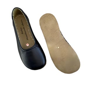 May include: Black leather ballet flats with a rounded toe and a separate sole. The shoe has the text "Swiss Handmade shoes" printed on the insole. The sole is a light brown color.