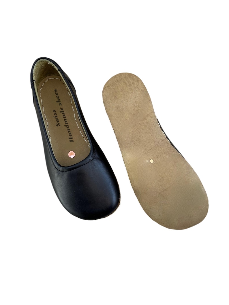 May include: Black leather ballet flats with a rounded toe and a separate sole. The shoe has the text "Swiss Handmade shoes" printed on the insole. The sole is a light brown color.