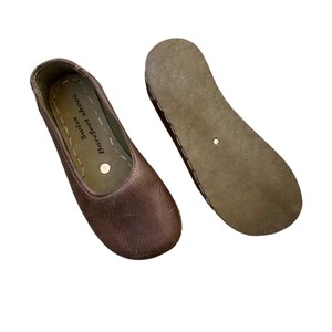 May include: Brown leather Swiss Barefoot Shoes, a pair of minimalist flats. One shoe is right-side up, displaying the upper with the brand name. The other shoe is upside down, showing the sole. The shoes are designed for comfort and natural movement.
