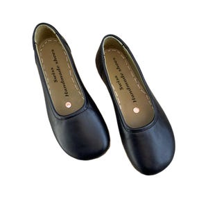May include: A pair of black leather ballet flats. The shoes have a rounded toe and a stitched sole. The insole is tan with the text "Swiss Handmade shoes". A small copper-colored dot is visible on the insole.