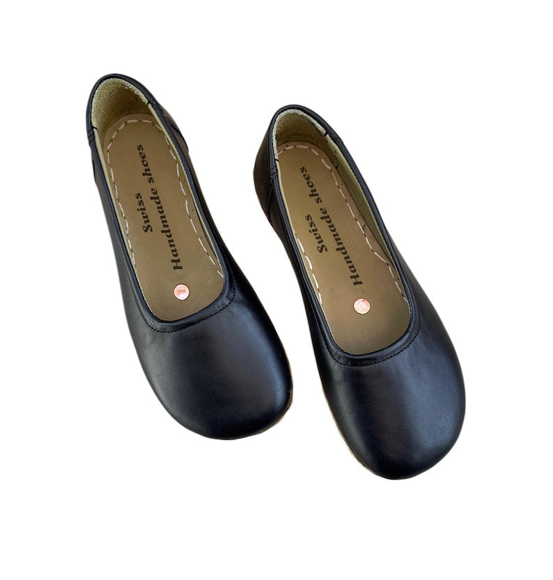 May include: A pair of black leather ballet flats. The shoes have a rounded toe and a stitched sole. The insole is tan with the text "Swiss Handmade shoes". A small copper-colored dot is visible on the insole.