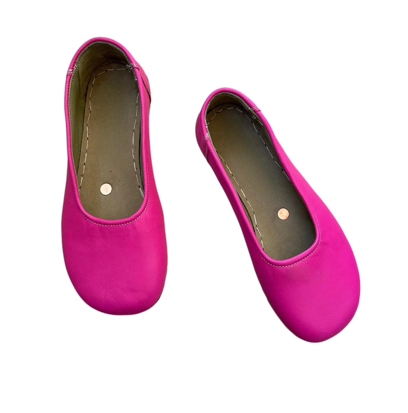 May include: A pair of bright pink ballet flats. The shoes have a rounded toe and a simple design. The interior is a neutral color, and the shoes are set against a white background. The shoes are made of a smooth material.