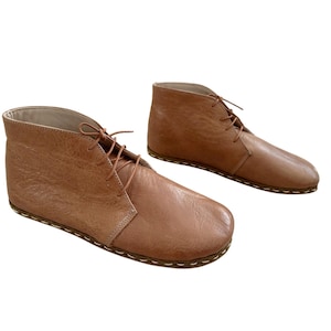 Könnte beinhalten: Ein Paar hellbraune Leder-Ankle-Boots mit Schnürsenkeln. Die Stiefel haben eine runde Spitze und eine genähte Sohle. Das Leder hat ein strukturiertes Aussehen. Die Stiefel sind vor einem weißen Hintergrund platziert.