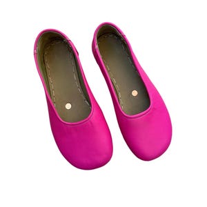 May include: A pair of bright pink ballet flats. The shoes have a rounded toe and a flat sole. The interior is a muted green color with a small, round, beige accent. The shoes are made of a smooth material.