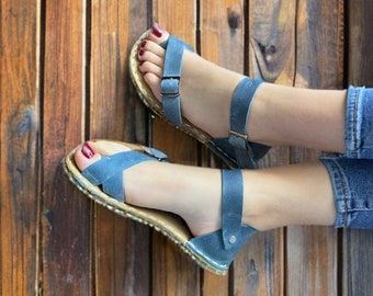 May include: A pair of blue leather sandals with a buckle closure. The sandals are worn on a person's feet with red nail polish on their toes.
