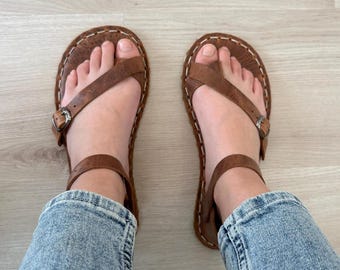 Puede incluir: Un par de sandalias de cuero marrón con cierre de hebilla. Las sandalias tienen una correa de estilo thong y se usan en los pies de una persona.