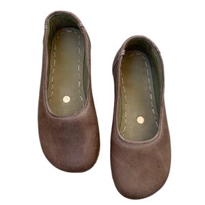May include: A pair of brown leather ballet flats. The shoes have a rounded toe, a stitched seam around the opening, and a small, round accent on the insole. The leather has a slightly distressed appearance.