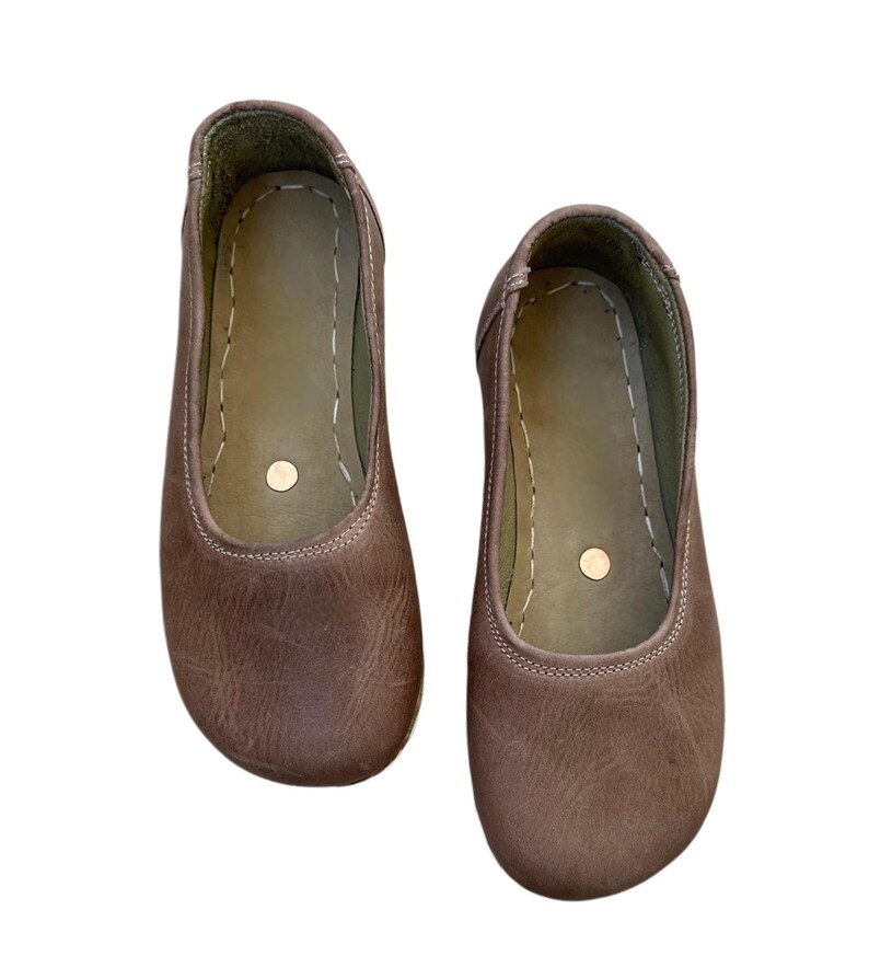 May include: A pair of brown leather ballet flats. The shoes have a rounded toe, a stitched seam around the opening, and a small, round accent on the insole. The leather has a slightly distressed appearance.